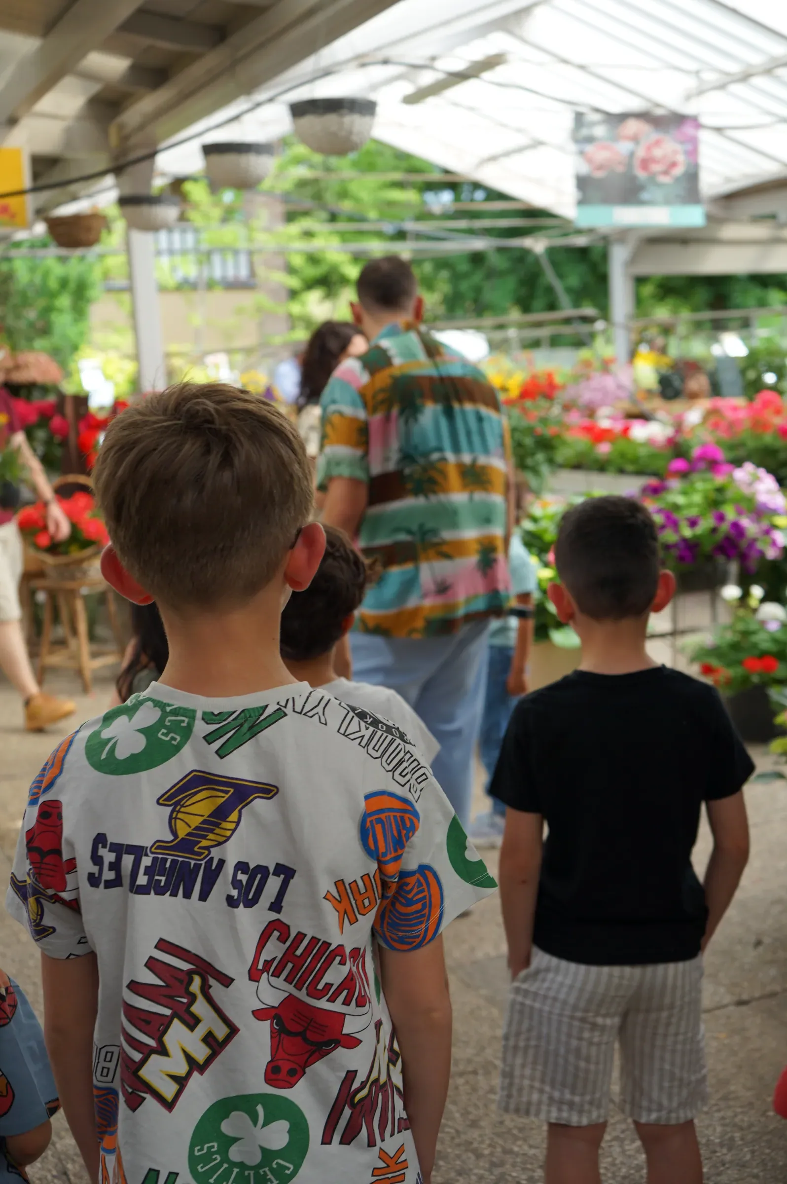 Due bambini di spalle fra i banchi di fiori sotto la copertura del garden, con un adulto in camicia a fantasia