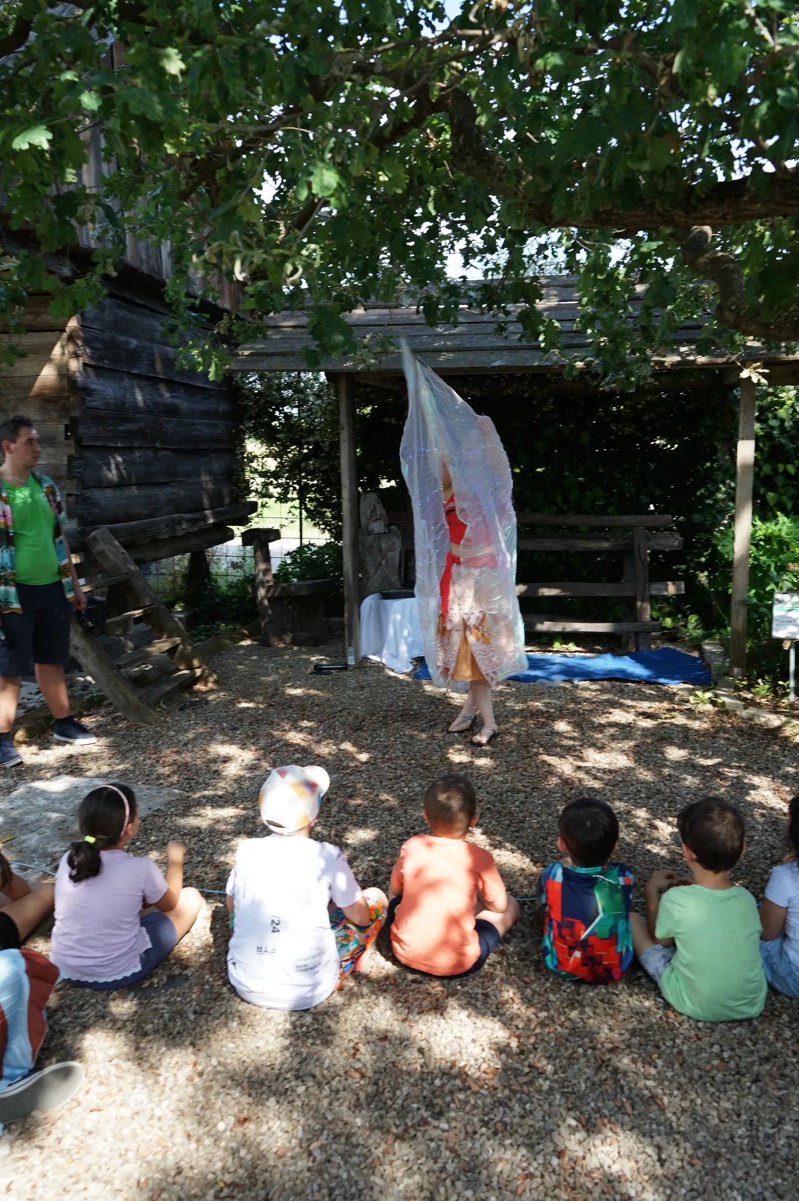 Bambini seduti di spalle sulla ghiaia guardano Vaiana, nascosta dietro un grande velo trasparente sotto la tettoia di legno