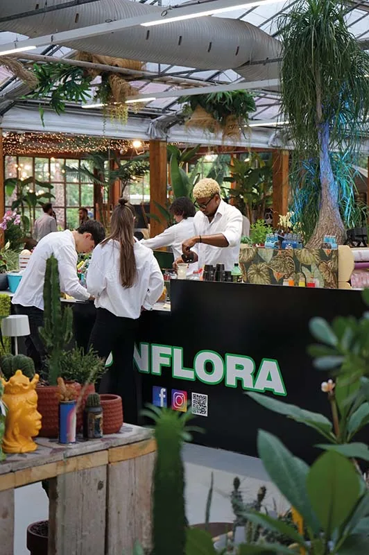 Il bancone nero con la scritta Sinflora sotto la serra, dove i baristi in camicia bianca preparano i cocktail fra cactus e piante tropicali