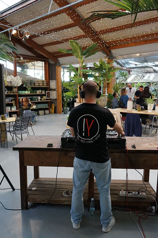 Il DJ di spalle alla consolle su un tavolo da lavoro in legno, sotto le travi e le lucine della serra, con gli ospiti ai tavoli sul fondo