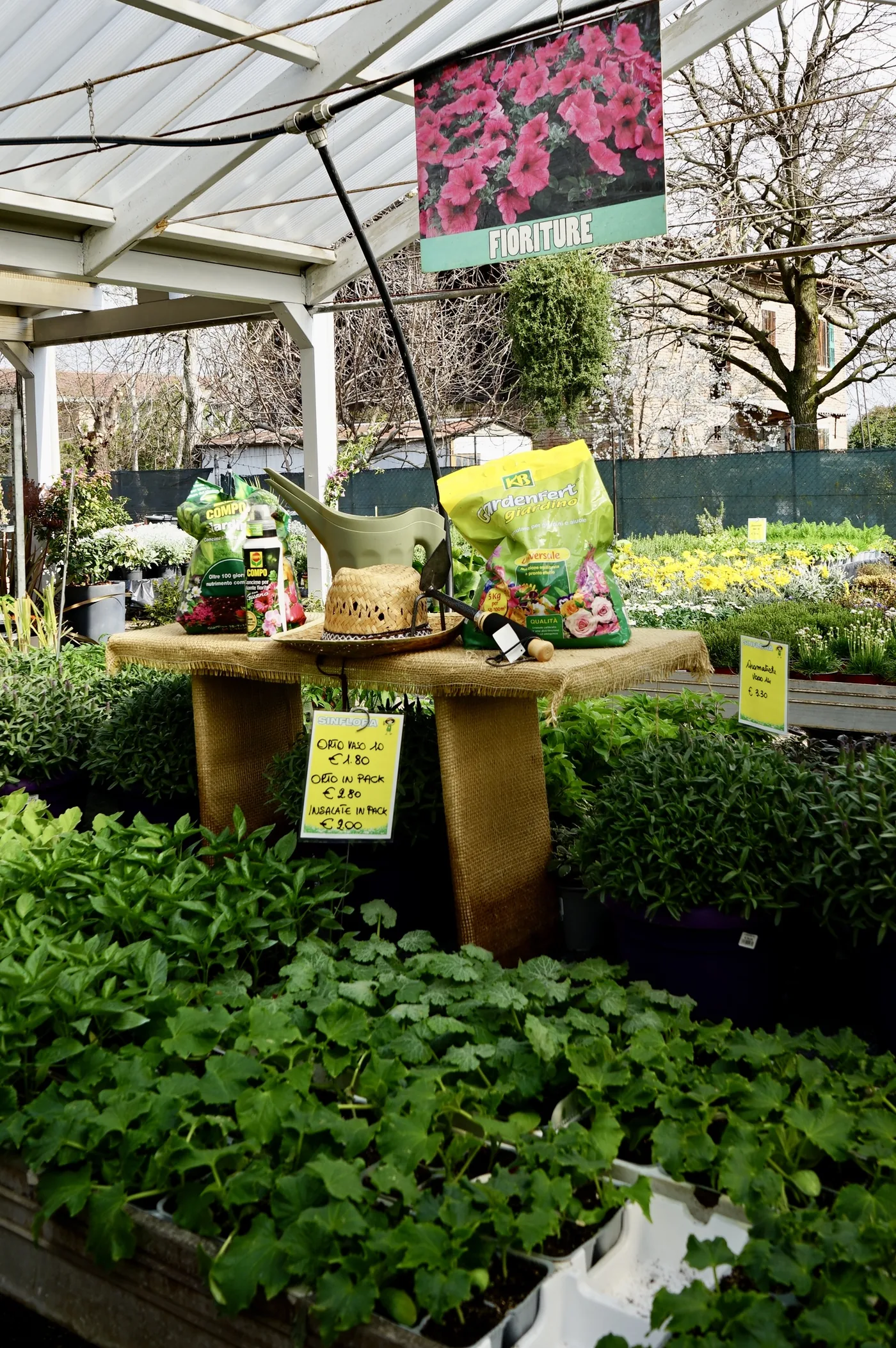 Banco di piantine da orto e aromatiche sotto la tettoia del garden, con il cartello delle fioriture e un tavolino allestito