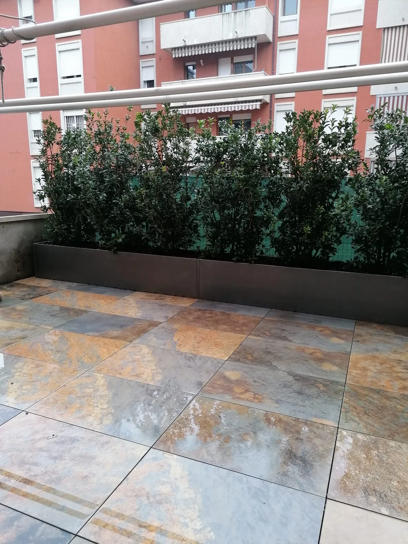 Terrazzo con lastre dai toni ruggine e grigi, fioriere in metallo e una siepe alta lungo il parapetto