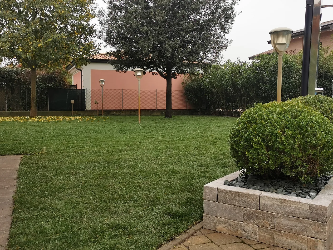 Prato con un'aiuola rialzata in blocchi di pietra e una grande sfera di bosso, alberi e case sul fondo