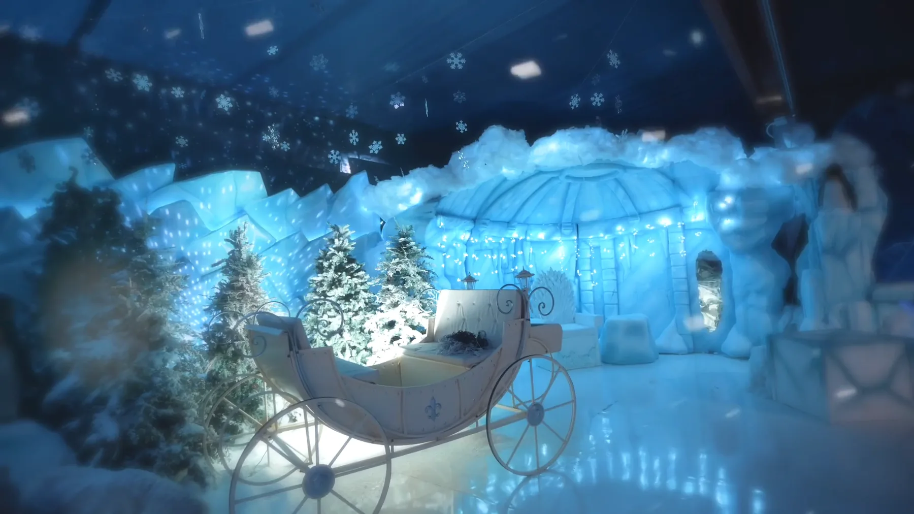 Una slitta bianca illuminata fra abeti innevati, davanti a una scenografia di ghiaccio azzurro con un igloo sotto un cielo di fiocchi di neve