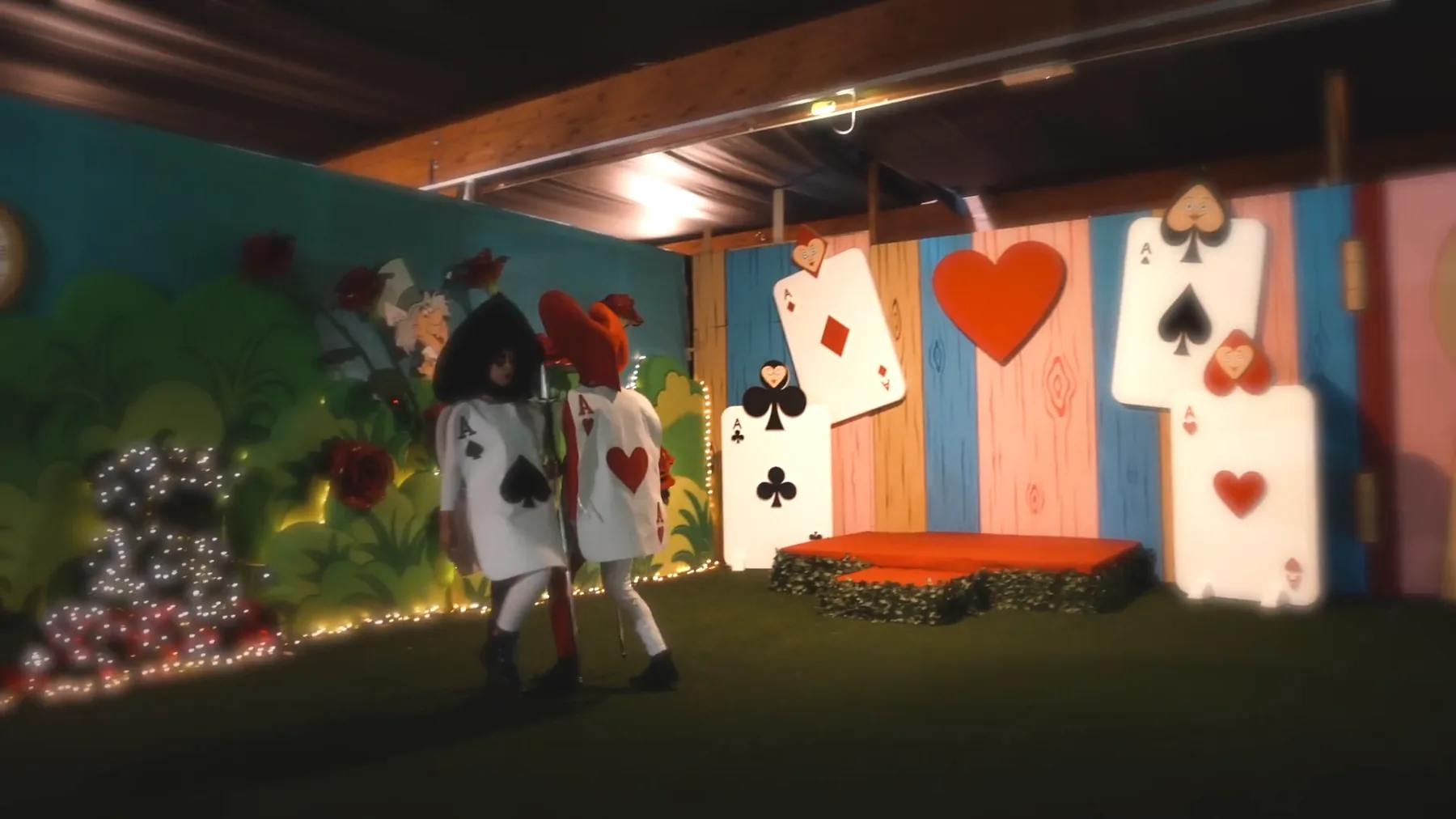 Scenografia con grandi carte da gioco appese, un cuore rosso, un piccolo palco e due figuranti vestiti da carte