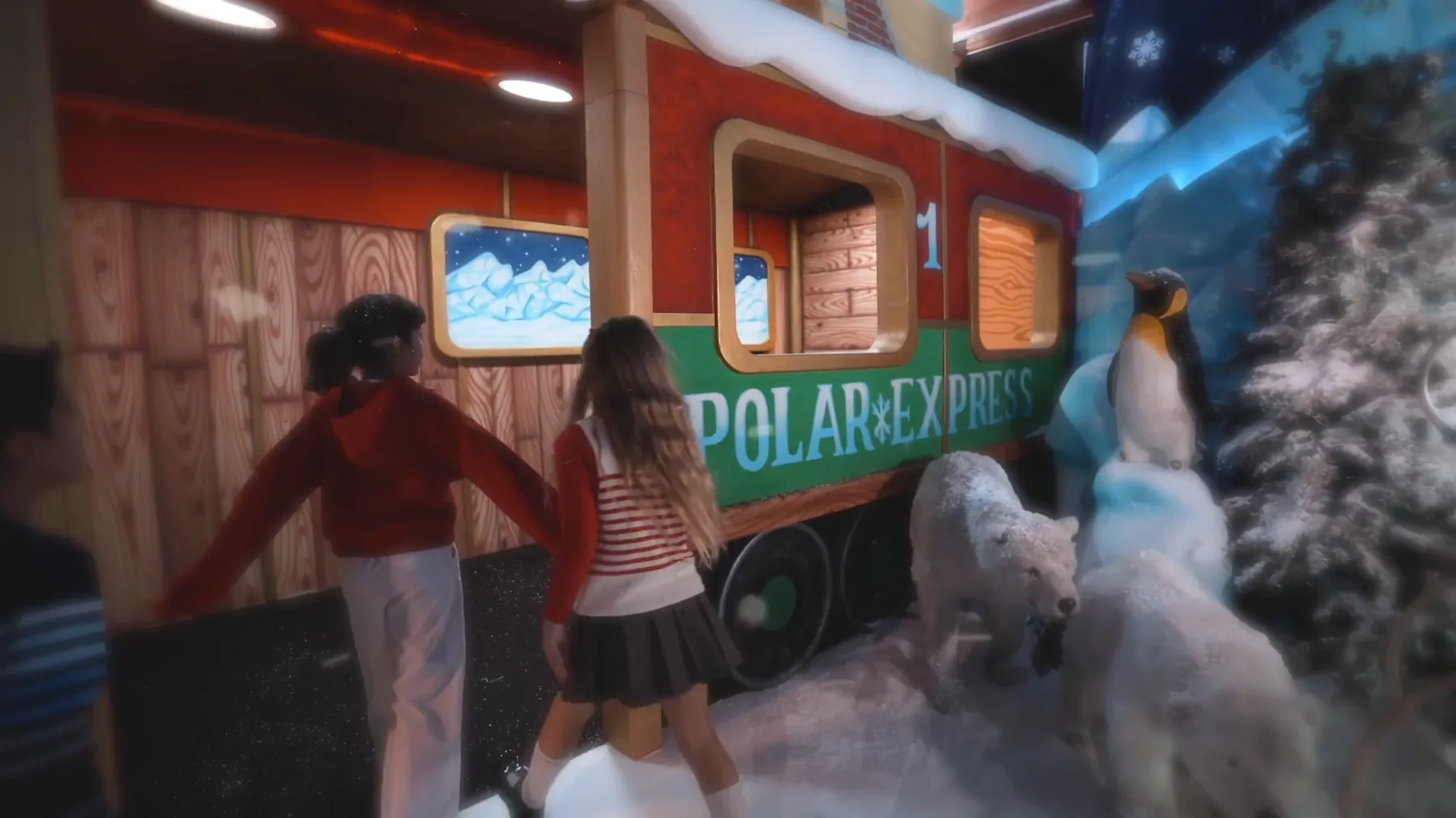 Due bambine di spalle davanti a un vagone rosso e verde con la scritta Polar Express, fra orsi polari e un pinguino