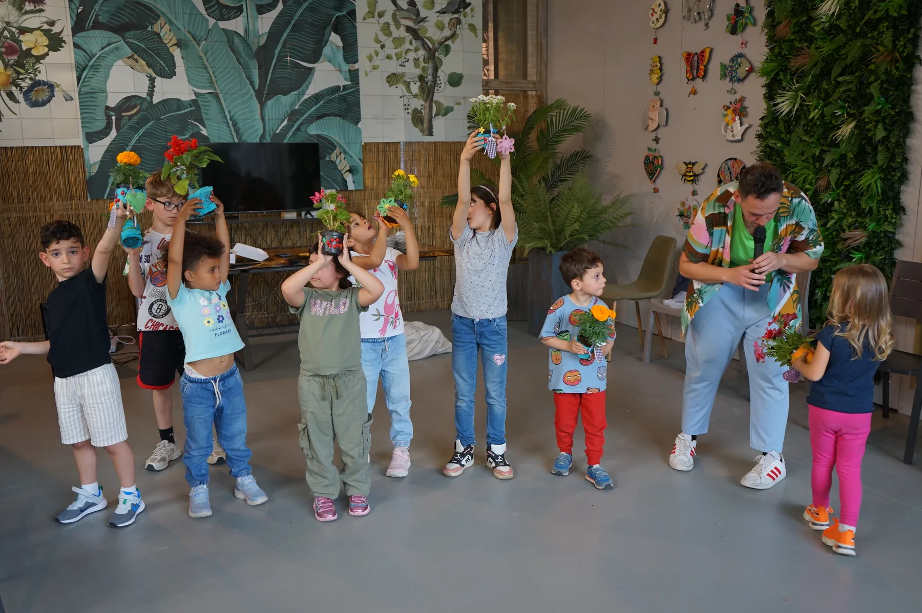 Bambini in fila sul pavimento alzano sopra la testa i vasetti decorati con le piantine, accanto a un animatore