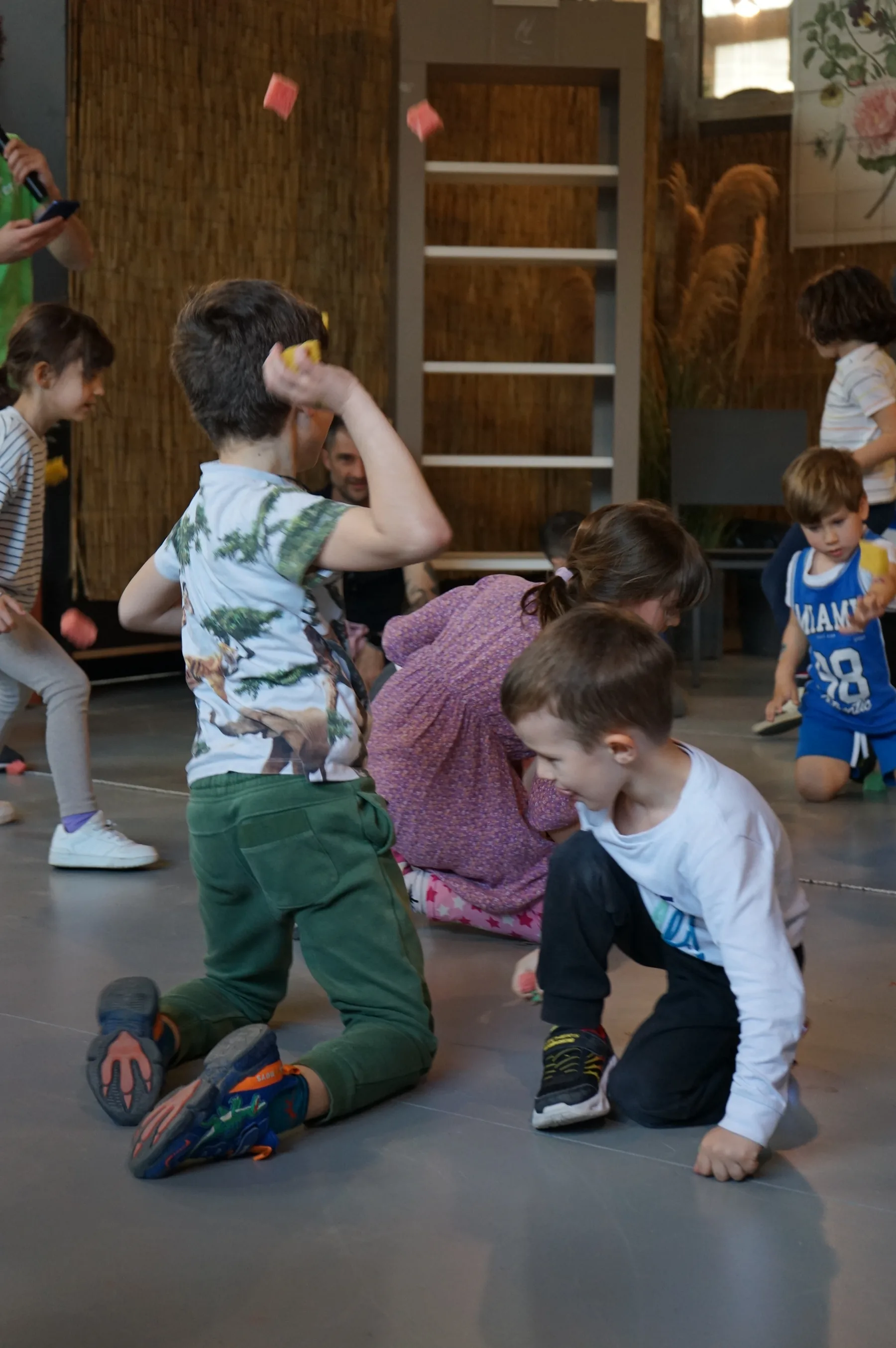 Bambini inginocchiati sul pavimento grigio che si lanciano piccoli cartoncini colorati, davanti a una parete di canne
