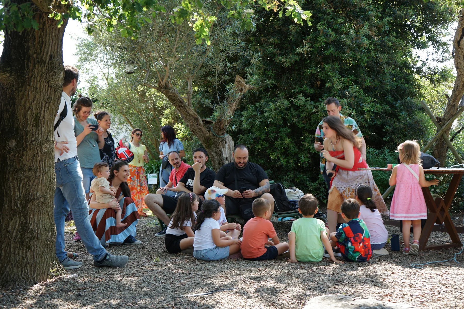 Bambini seduti sulla ghiaia all'ombra degli alberi, con i genitori attorno e Vaiana in piedi sulla destra