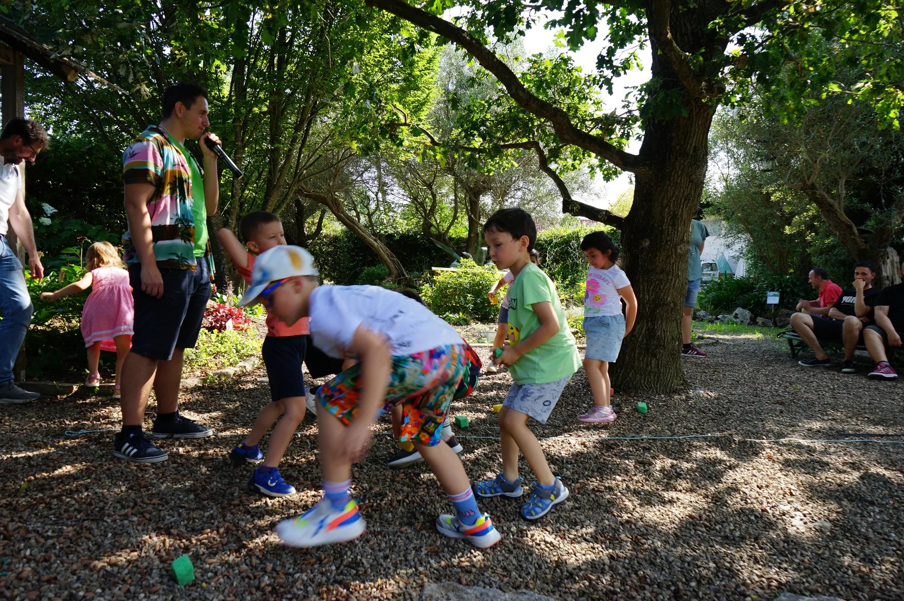 Un animatore con il microfono guida un gioco: i bambini chinati corrono fra i bicchierini colorati sulla ghiaia