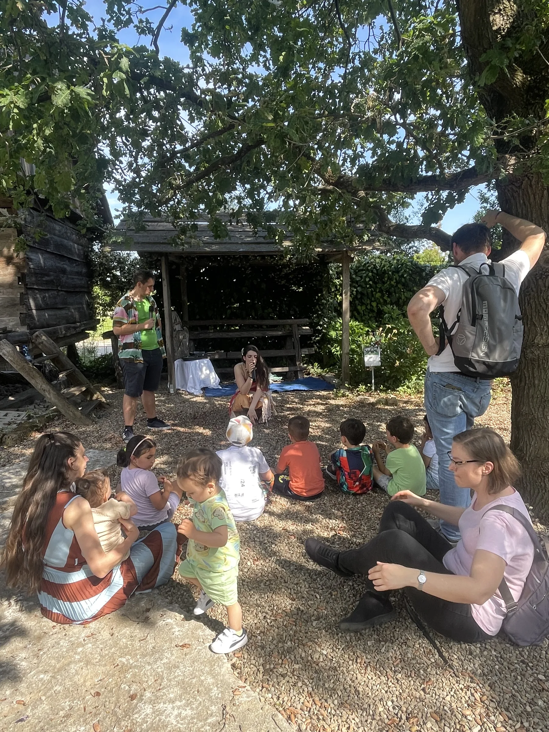 Bambini seduti per terra davanti alla capanna di legno, con i genitori seduti sulla ghiaia accanto a loro