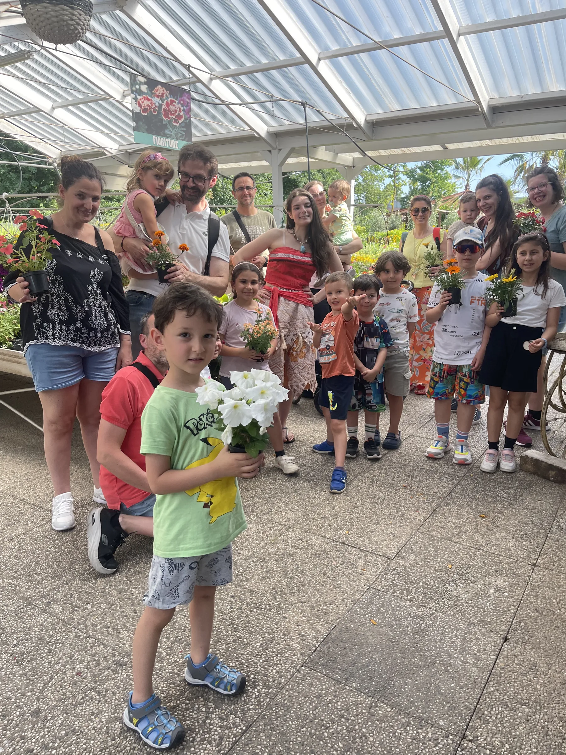 Bambini e genitori sotto la tettoia del garden con Vaiana in mezzo; in primo piano un bambino tiene un vaso di fiori bianchi
