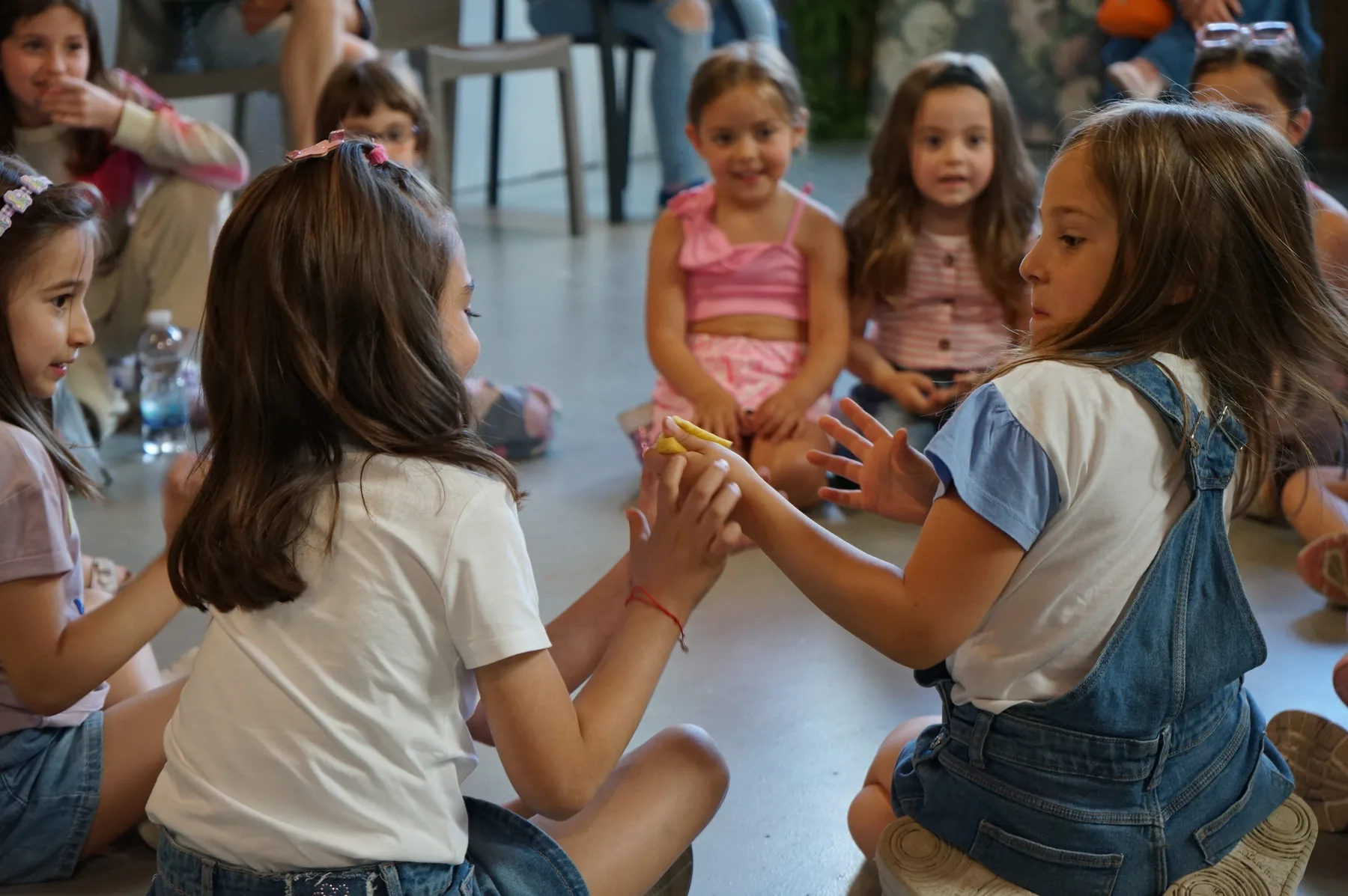 Due bambine sedute una di fronte all'altra si passano qualcosa fra le mani, mentre le altre guardano