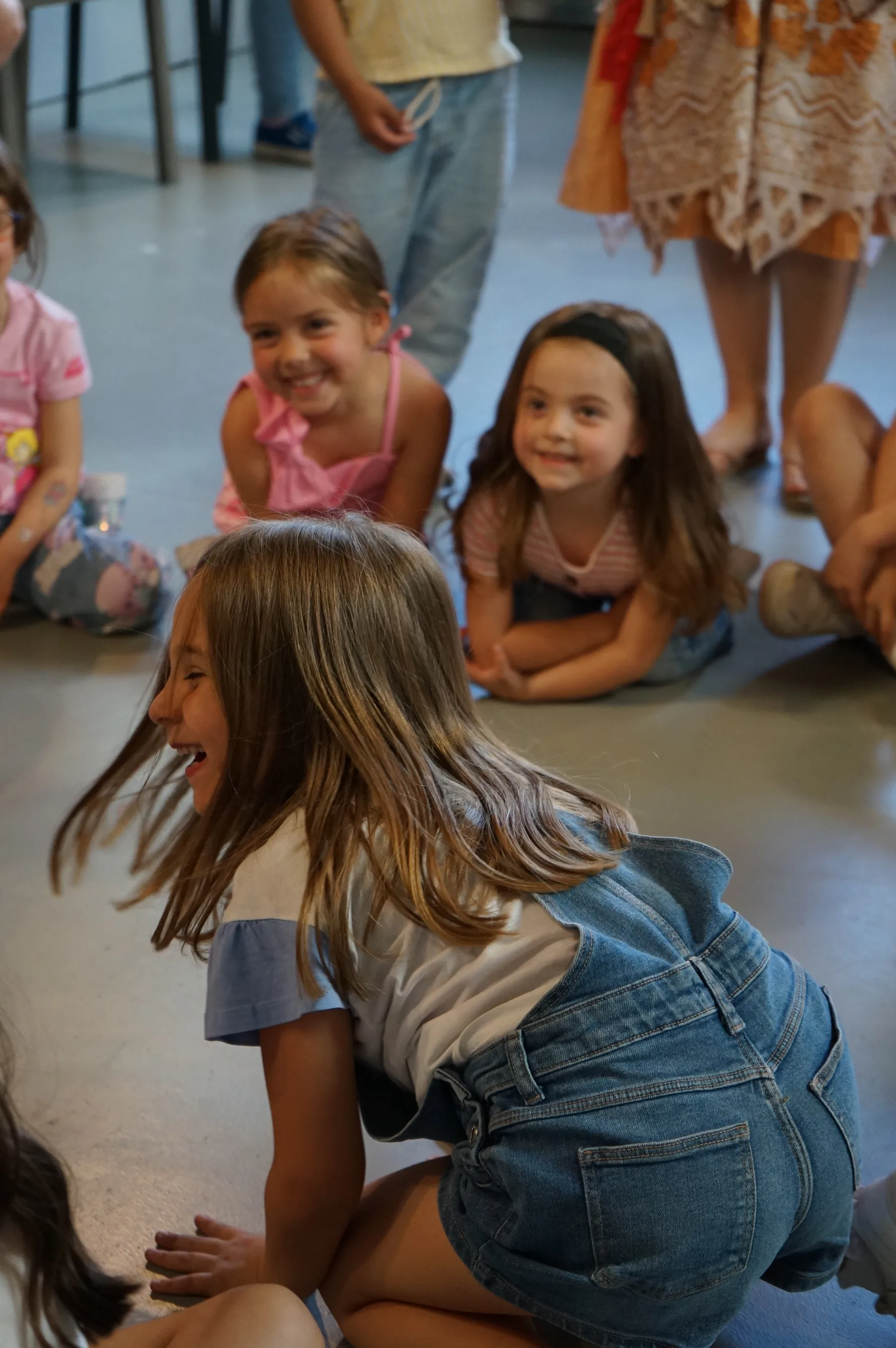 Una bambina con la salopette di jeans si gira ridendo, seduta sul pavimento accanto ad altre due bambine