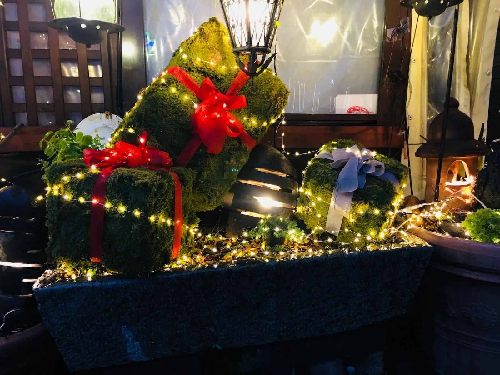 Fioriera con pacchi regalo di muschio legati con nastri rossi e lucine, davanti a un locale
