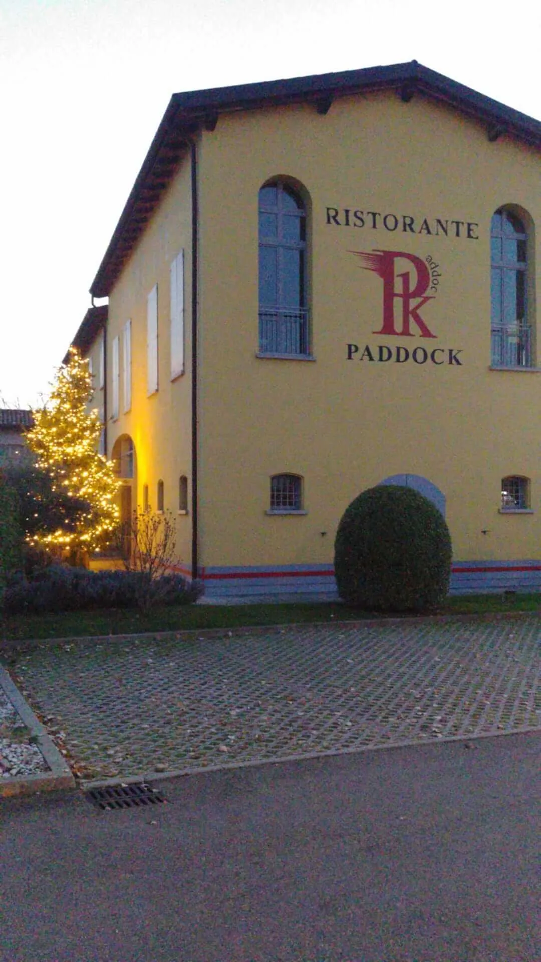 Facciata gialla del ristorante Paddock con un albero illuminato accanto all'ingresso