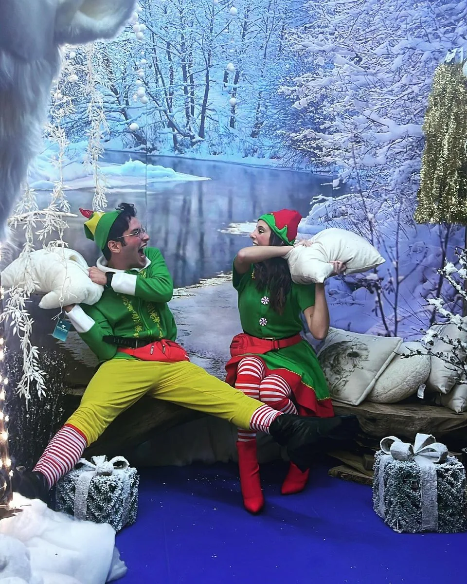 Due elfi in costume verde e rosso fanno la lotta con i cuscini davanti a un paesaggio innevato