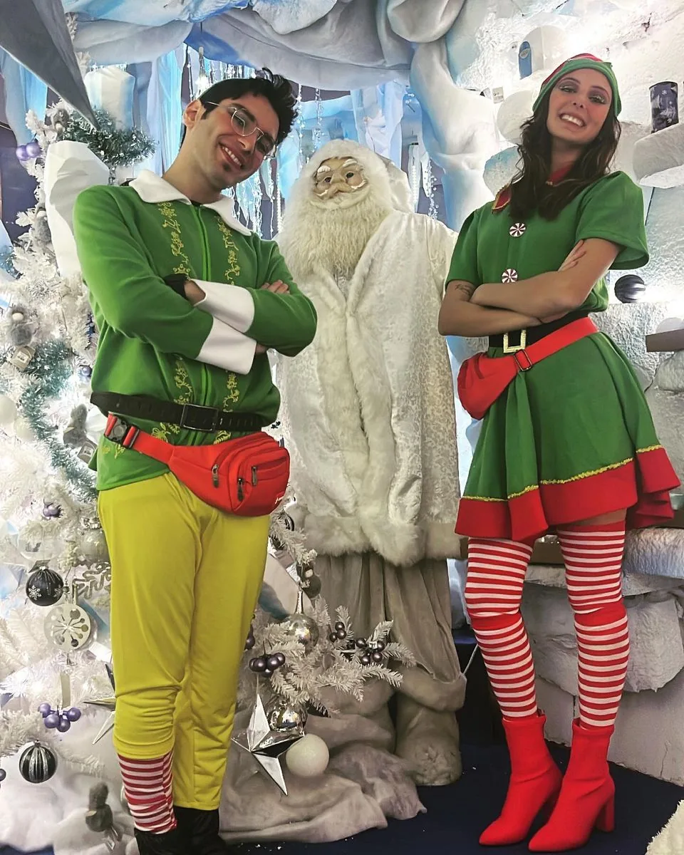 Due elfi a braccia conserte ai lati di un Babbo Natale bianco, davanti a un albero addobbato