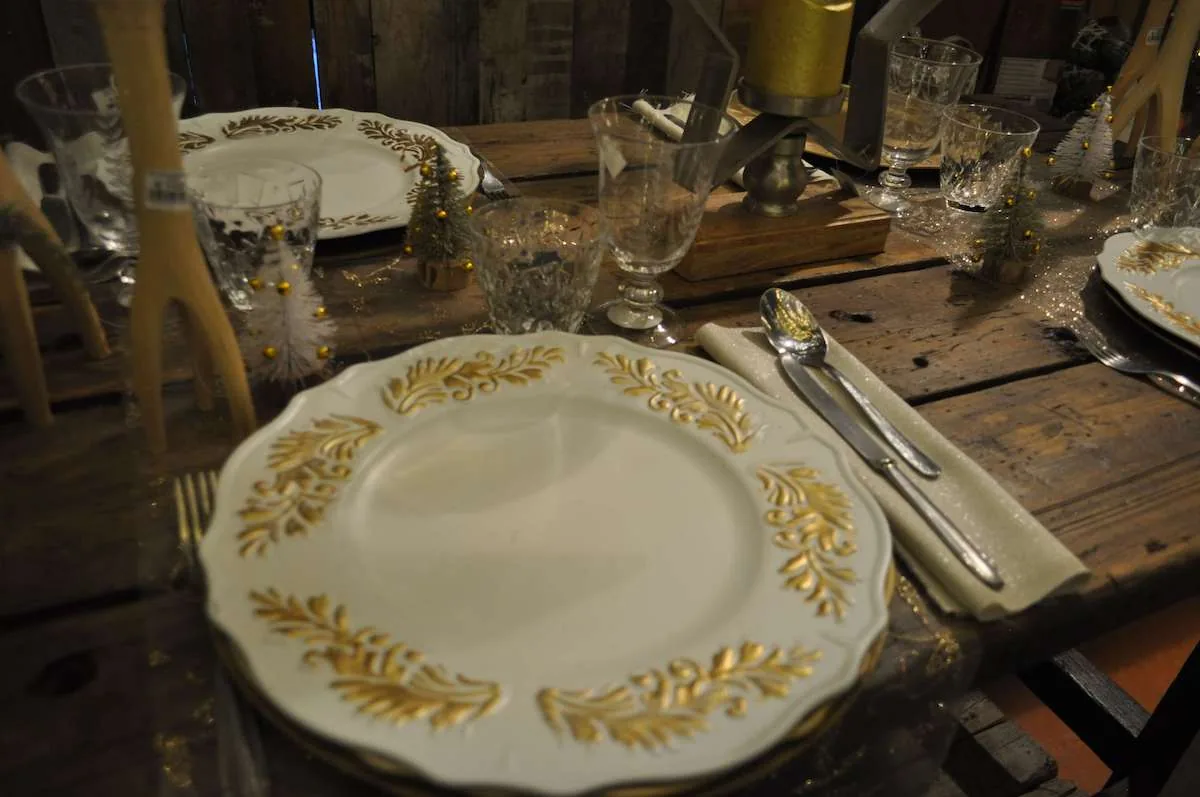 Piatto bianco con decori dorati apparecchiato su un tavolo di legno, con bicchieri di cristallo