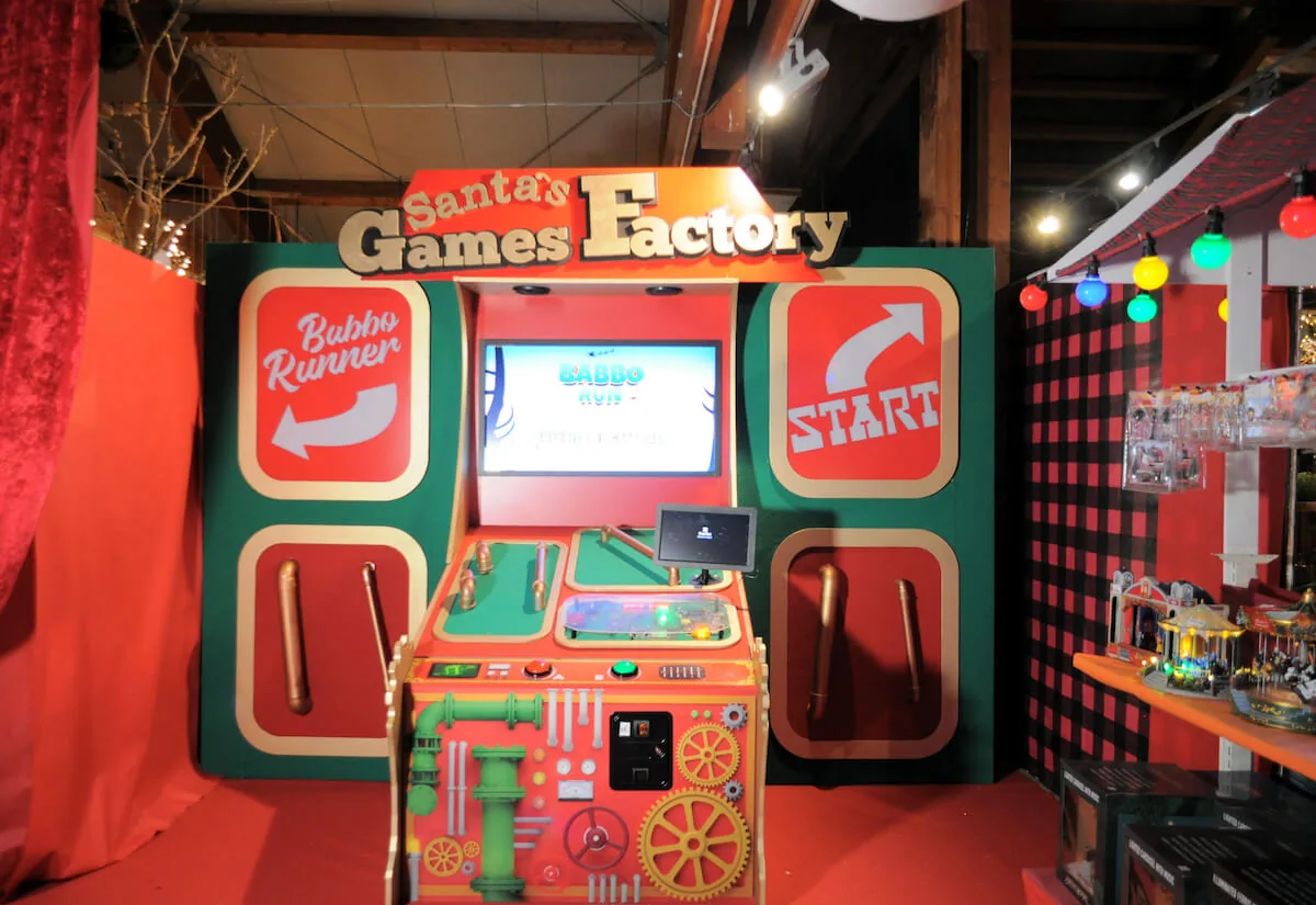 Postazione di gioco Santa's Games Factory, rossa e verde, con uno schermo e il pulsante di partenza