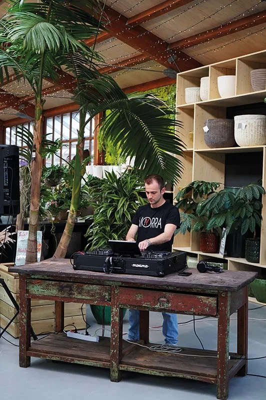 Il DJ alla consolle, appoggiata su un vecchio tavolo da lavoro in legno dipinto, fra le palme e gli scaffali dei vasi sotto la serra