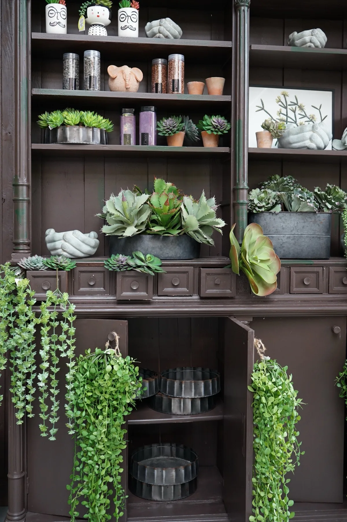 Credenza scura con succulente artificiali in vaschette di zinco, vasi a forma di mani in cemento e barattoli di ghiaia colorata