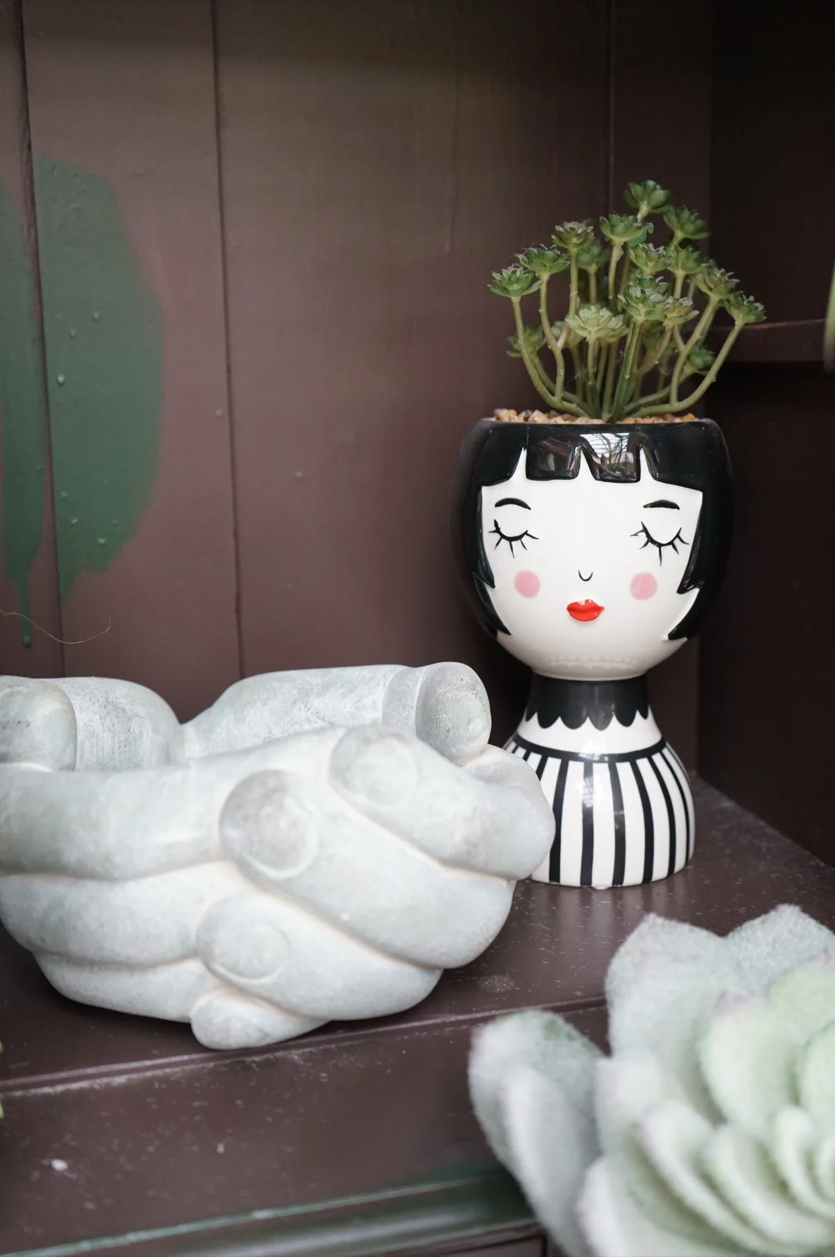 Vaso in ceramica bianca e nera a forma di volto con gli occhi chiusi, con una succulenta al posto dei capelli, accanto a una ciotola a forma di mani