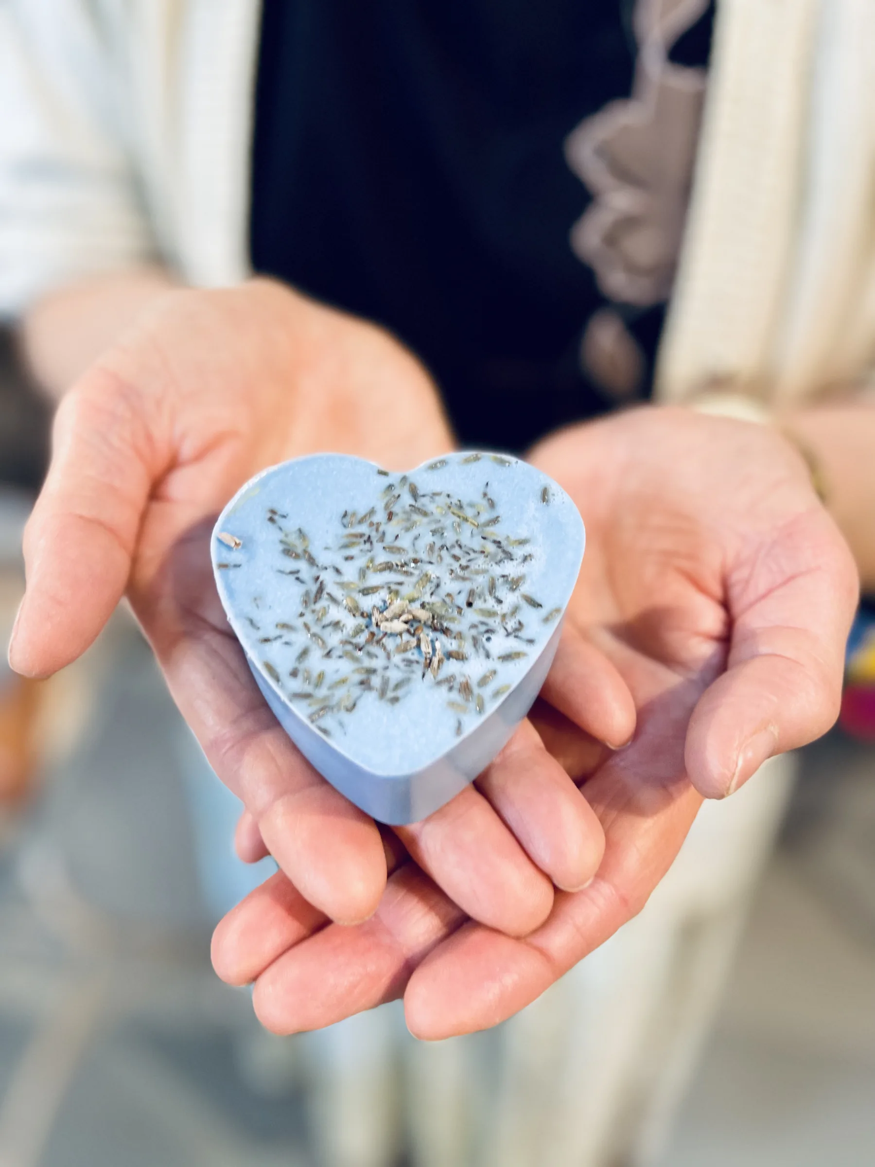 Due mani tengono un sapone azzurro a forma di cuore con i fiori di lavanda