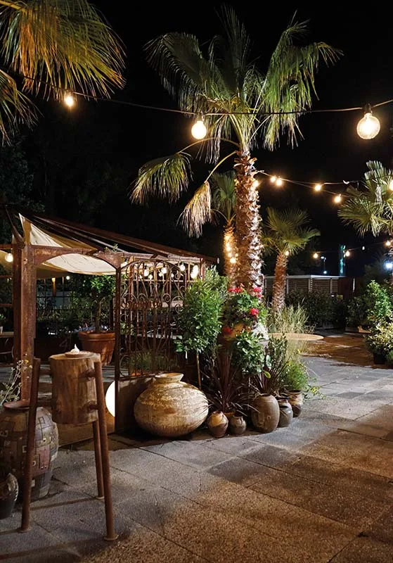 Il pergolato all'aperto di sera, con le lampadine accese fra le palme e le anfore di terracotta