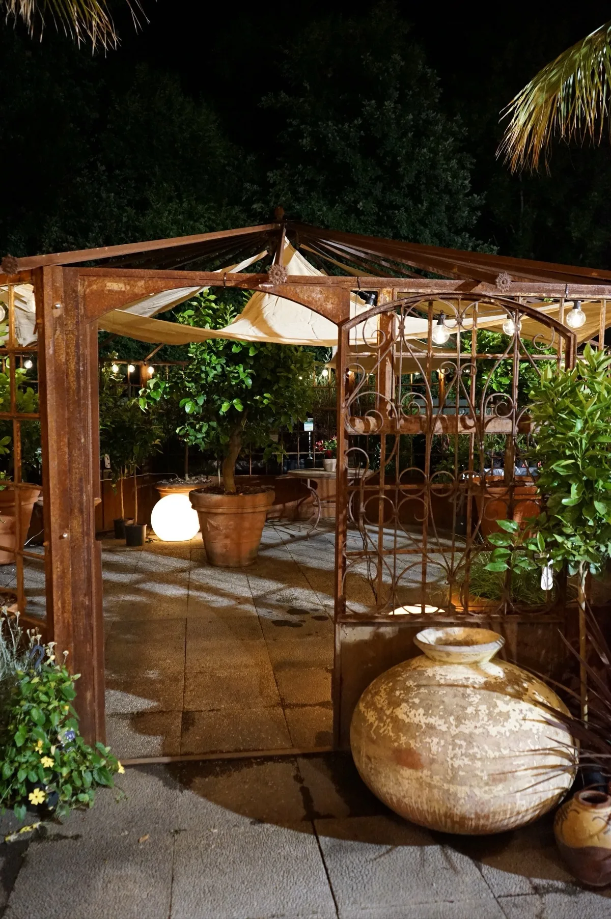 Il pergolato all'aperto di sera, con le lampadine accese fra le palme e un'anfora di terracotta