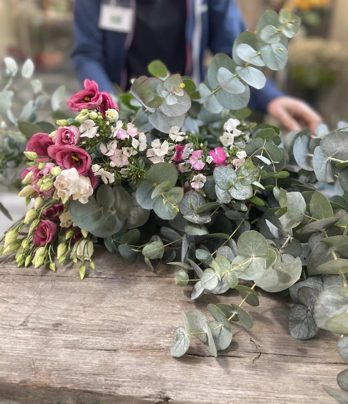 Lisianthus rosa e fiorellini bianchi appoggiati sul piano di legno del banco, in mezzo a rami di eucalipto, mentre la fiorista li dispone