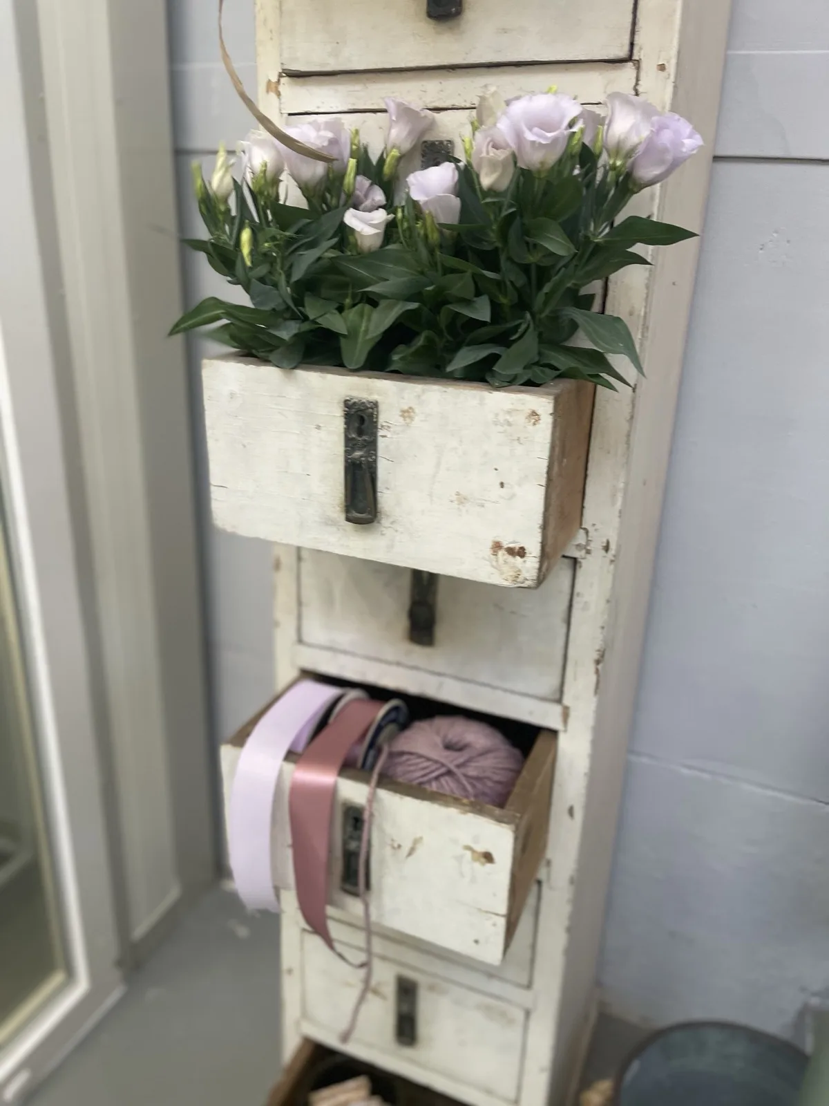 Cassettiera bianca scrostata con lisianthus lilla in un cassetto aperto e, in quello sotto, i rotoli di nastro rosa e un gomitolo di spago