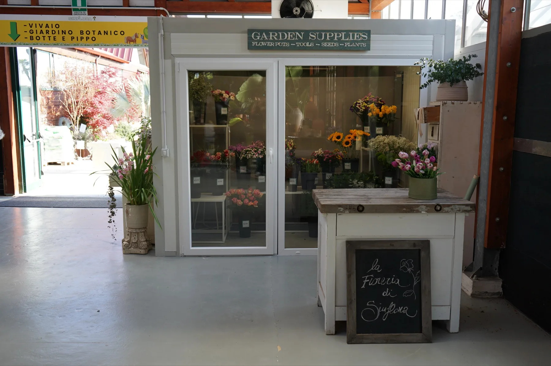 La cella frigo a vetri con dentro i fiori recisi, girasoli e tulipani, e accanto il banco della fioreria con la lavagna scritta a gesso