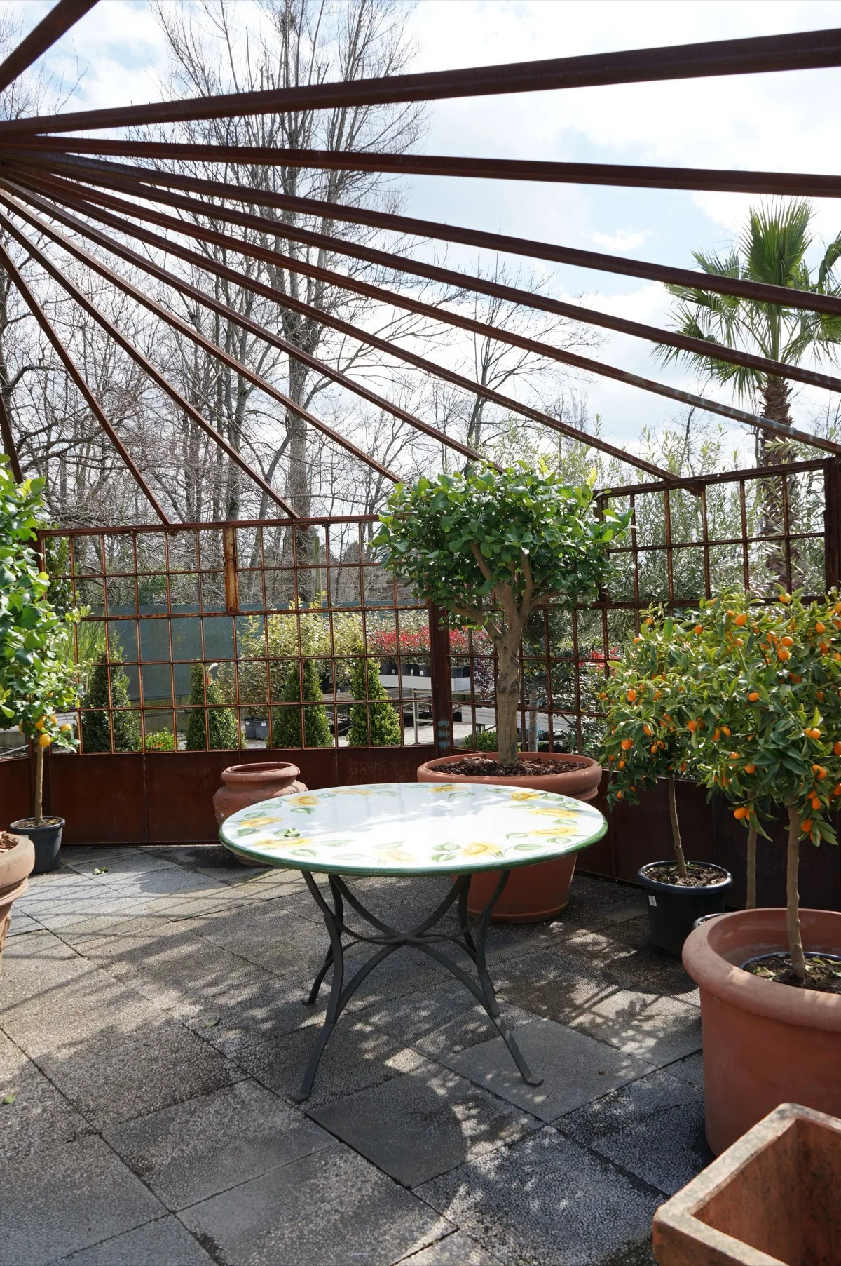 Sotto la struttura in ferro, un tavolo tondo in ceramica dipinta a limoni, circondato da limoni e kumquat in vasi di terracotta