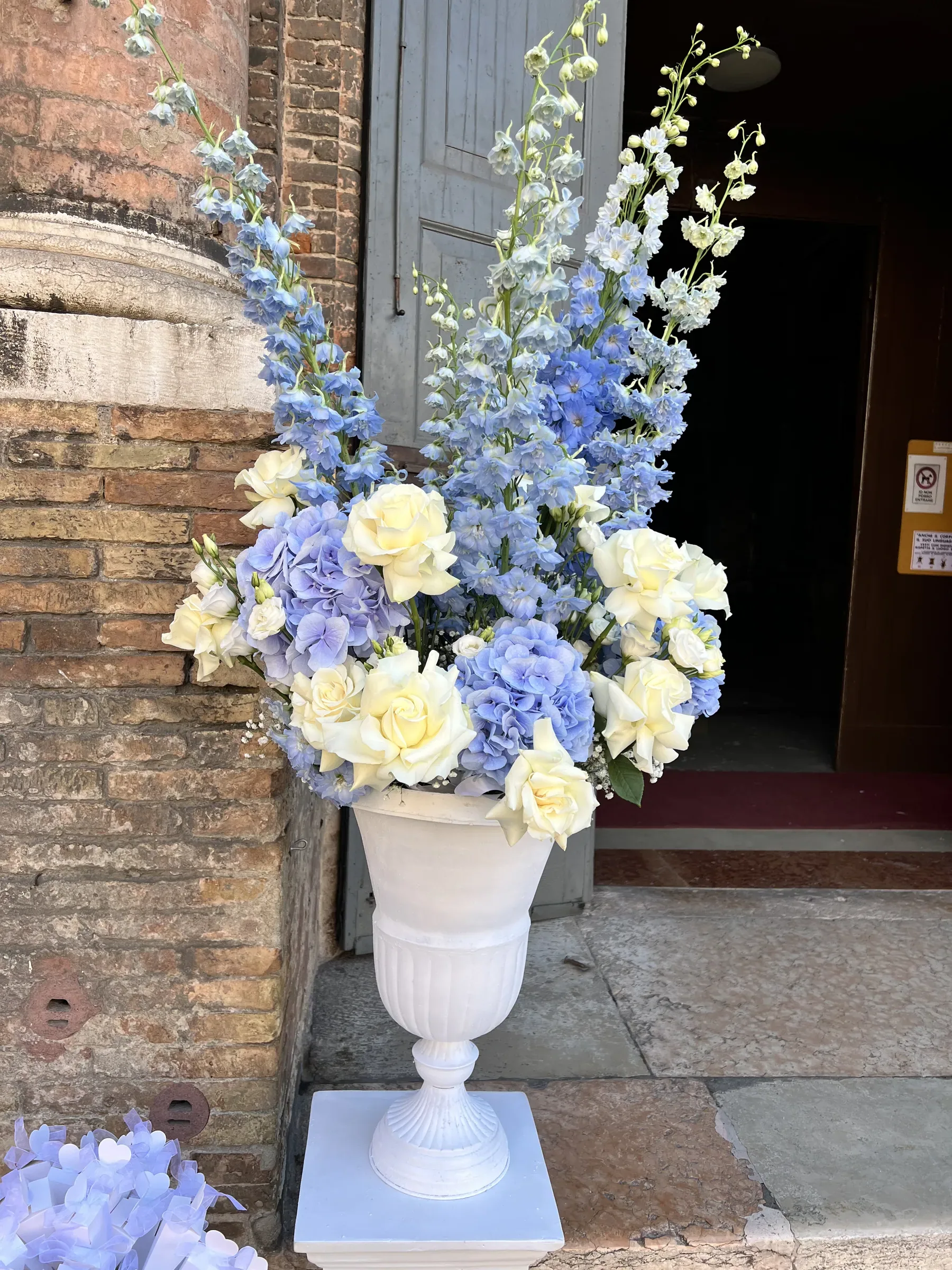 Composizione alta di delphinium azzurri e rose bianche in un'anfora bianca, davanti al portone di pietra di una chiesa