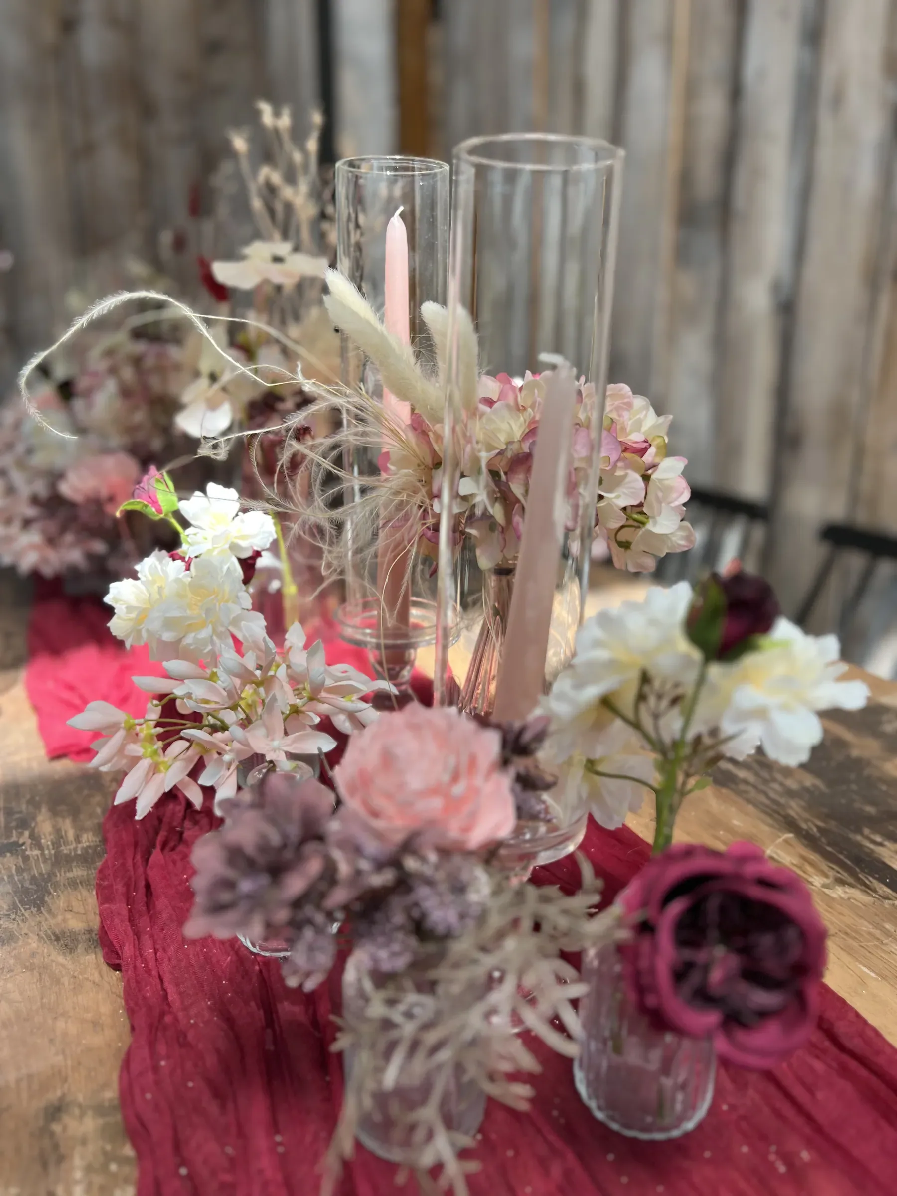 Centrotavola di fiori secchi e rose cipria con candele rosa in cilindri di vetro, su un runner bordeaux e un tavolo di legno