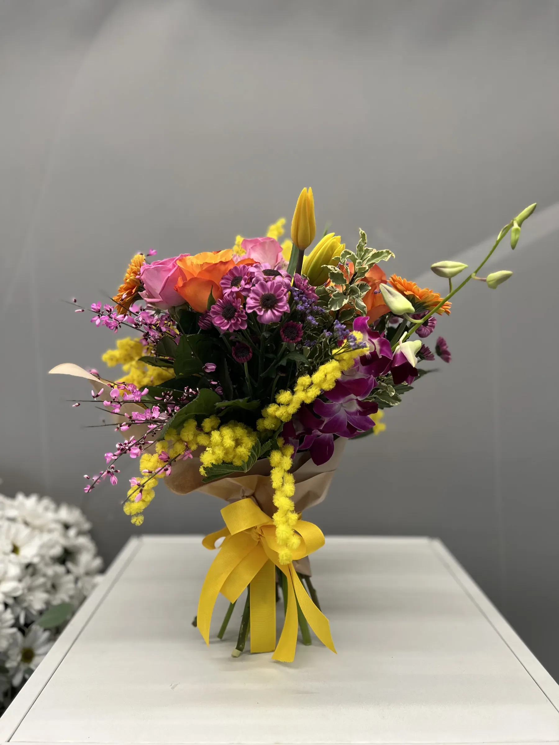 Mazzo di mimosa, gerbere e fiori viola legato con un nastro giallo, dritto su un piano bianco