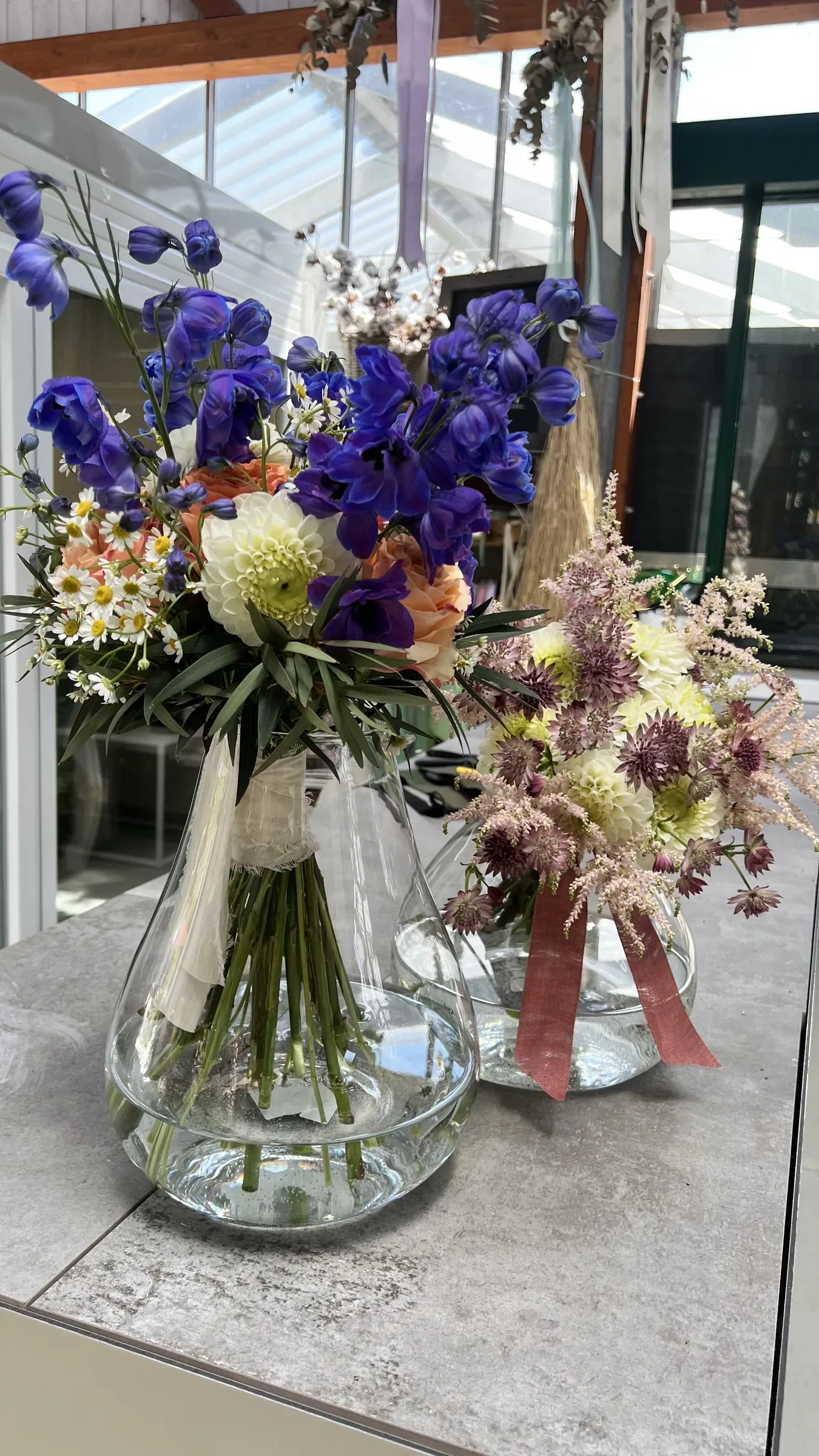Due vasi di vetro con delphinium blu, fiori bianchi e rosa pallido e nastri, su un piano di marmo sotto la serra