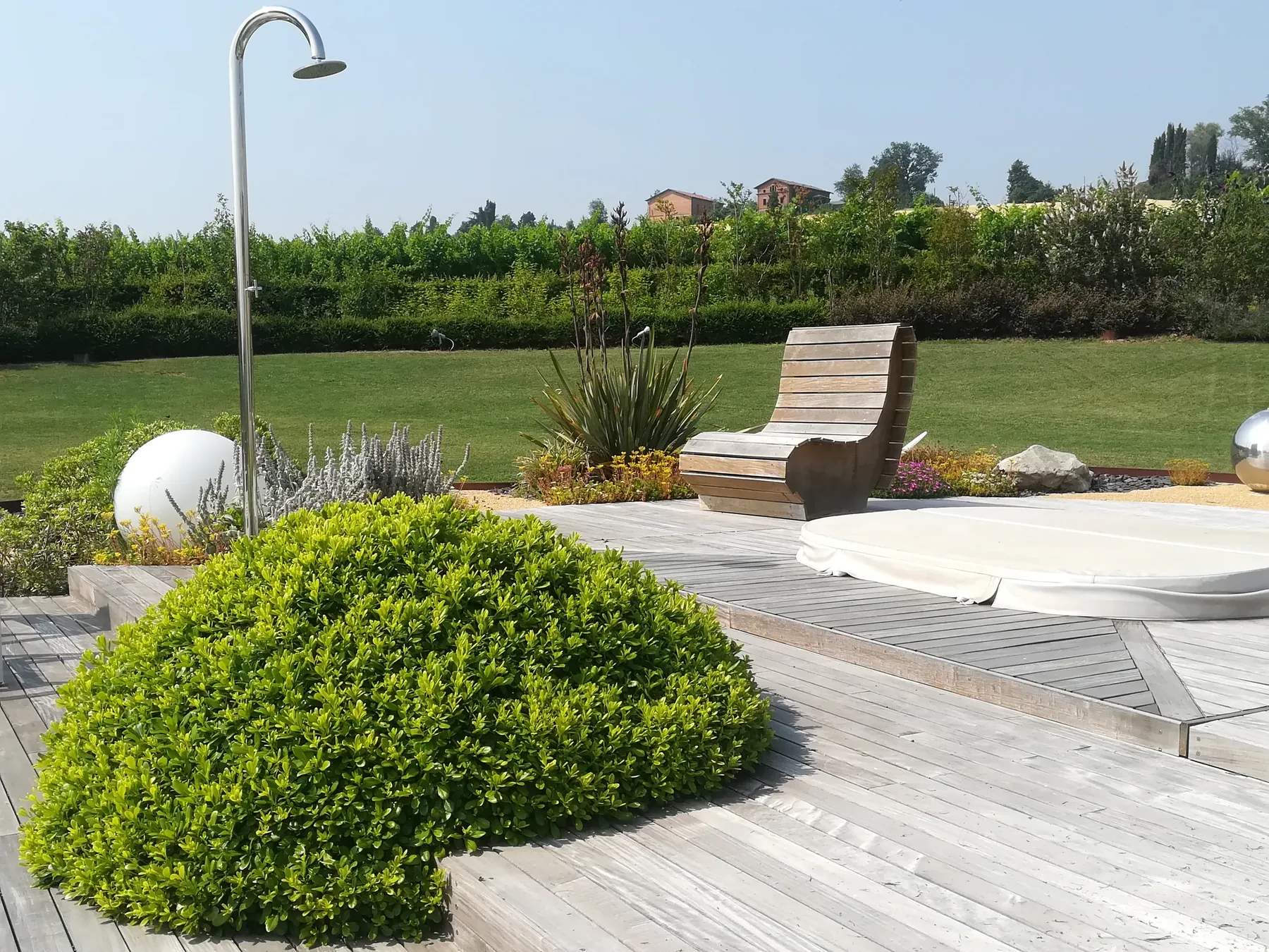 Cupola di bosso fra le doghe di una pedana in legno grigio, con una doccia da giardino, una sdraio e il prato dietro