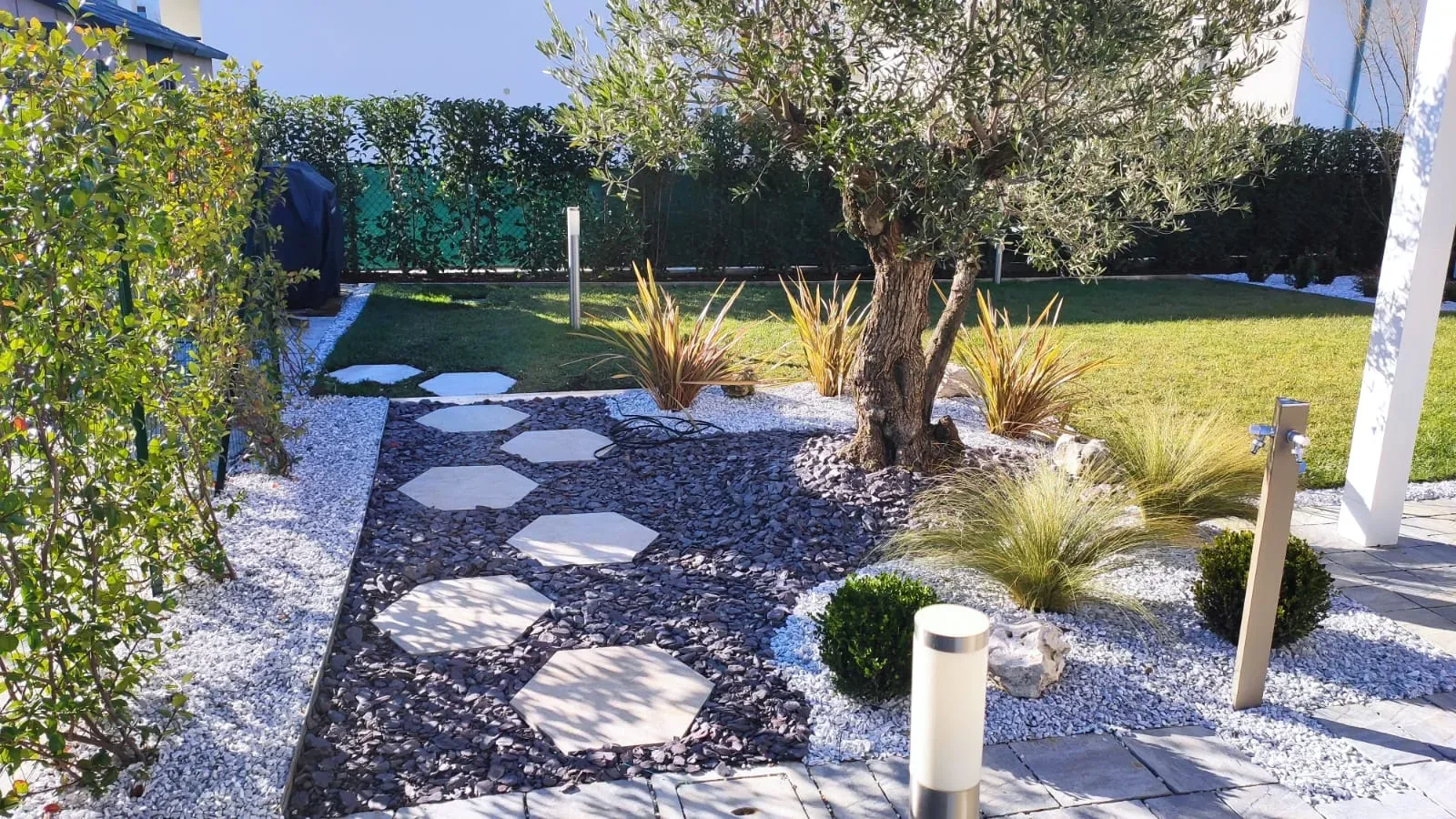 Passi esagonali in pietra chiara su ghiaia scura, bordata di ciottoli bianchi, con un olivo e graminacee lungo il prato