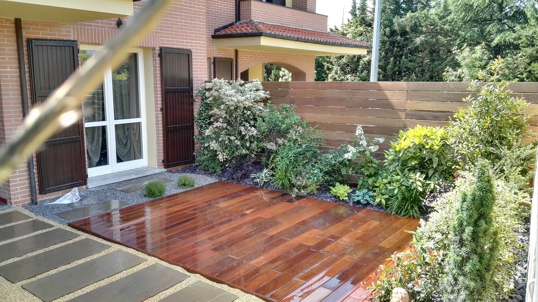 Pedana in legno fra lastre in pietra, con arbusti fioriti e un frangivista in legno lungo il confine