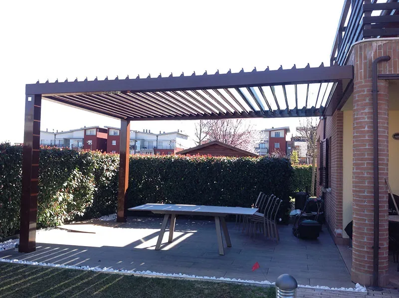 Pergola a lamelle color legno addossata a una casa in mattoni, sopra un tavolo lungo e una pavimentazione chiara
