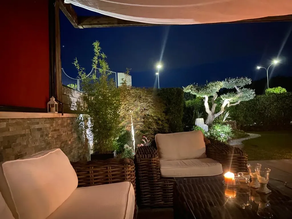 Salotto da esterno sotto la tenda, di sera, con le candele sul tavolino e un olivo illuminato in giardino