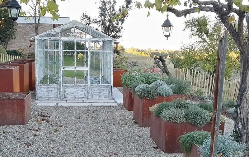 Piccola serra in ferro bianco su un cortile di ghiaia, fra fioriere alte in corten piene di piante argentate