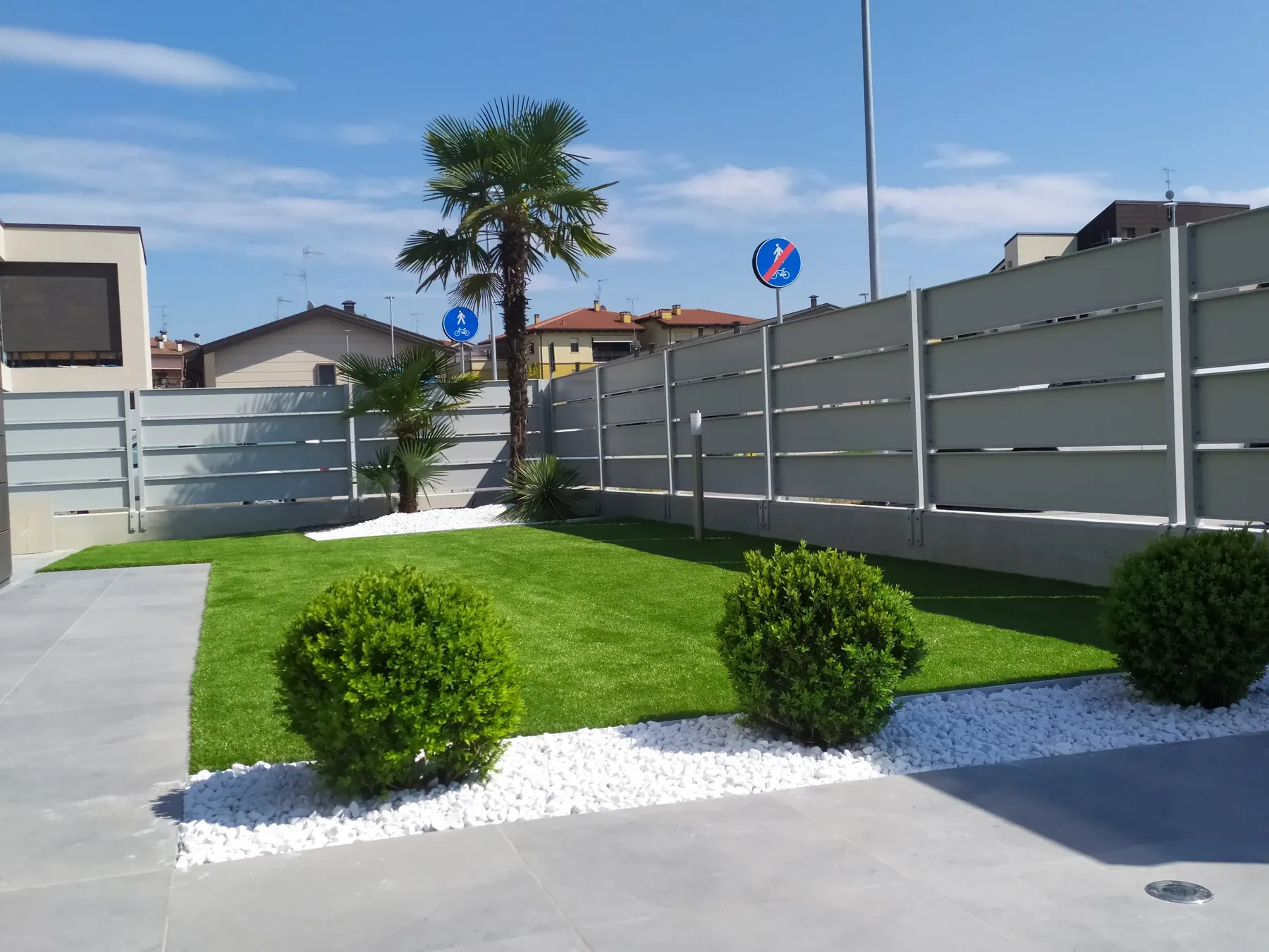Prato sintetico con tre sfere di bosso su ciottoli bianchi, una palma e la recinzione in doghe grigie