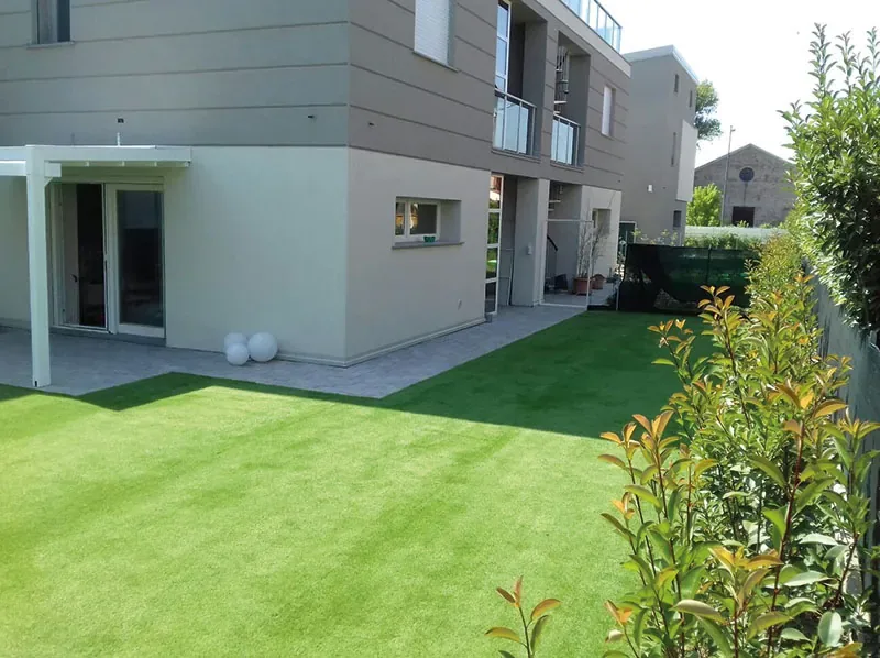 Prato sintetico davanti a una casa moderna, fra la pavimentazione e una fila di fotinie
