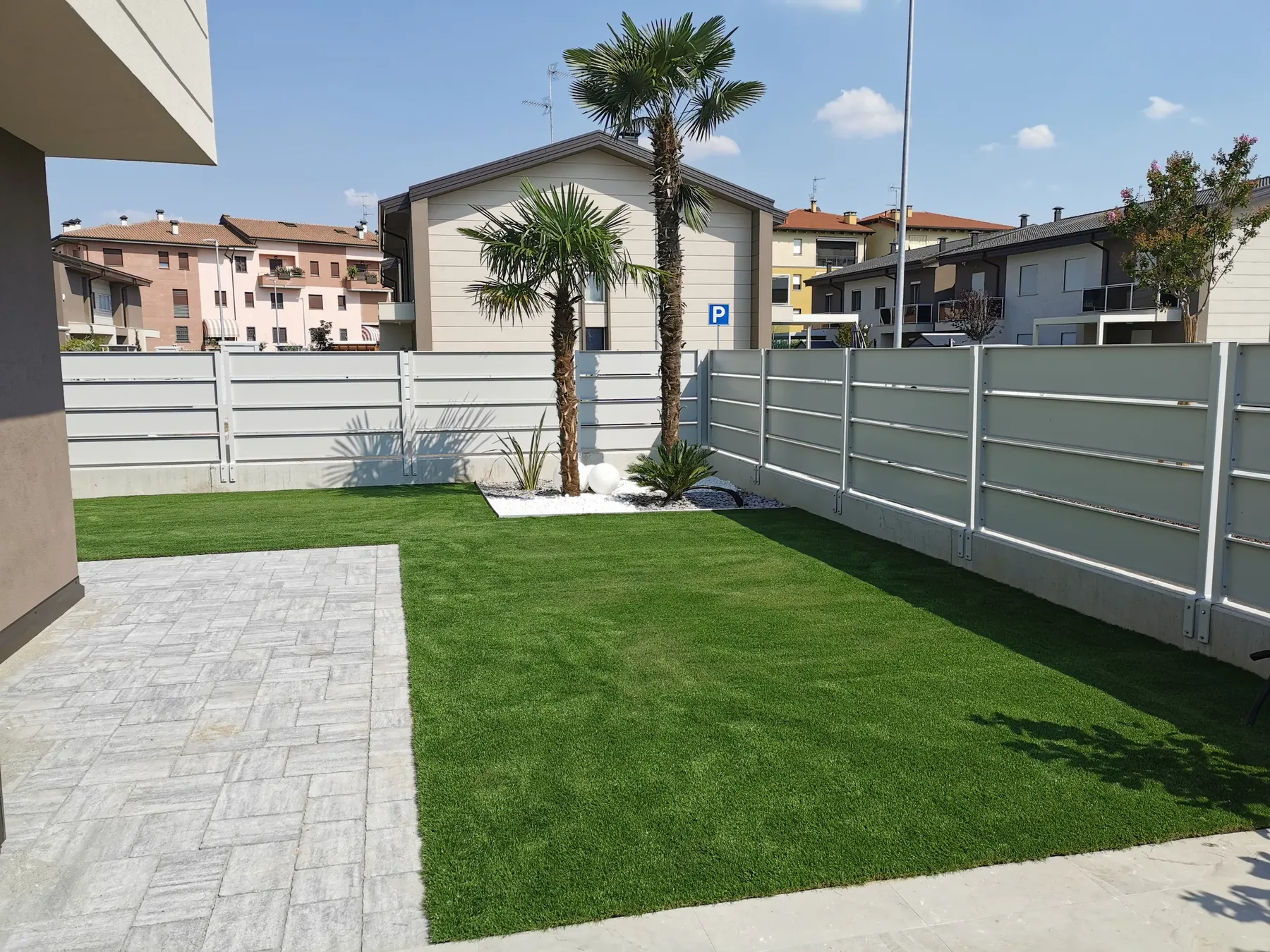 Prato sintetico con due palme su ghiaia bianca, fra il camminamento in pietra e la recinzione in doghe chiare