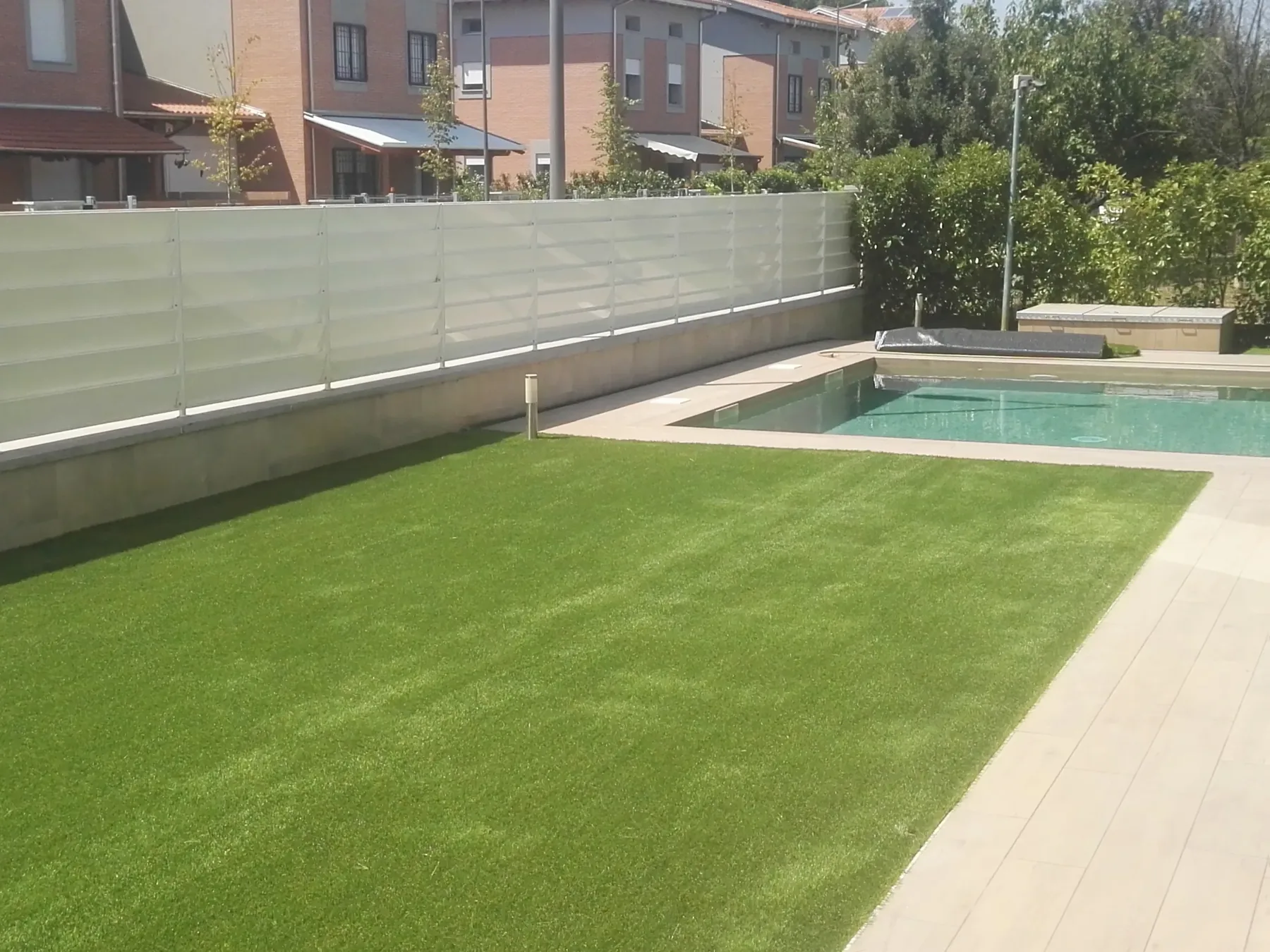 Prato sintetico fra il bordo in pietra chiara di una piscina e la recinzione bianca