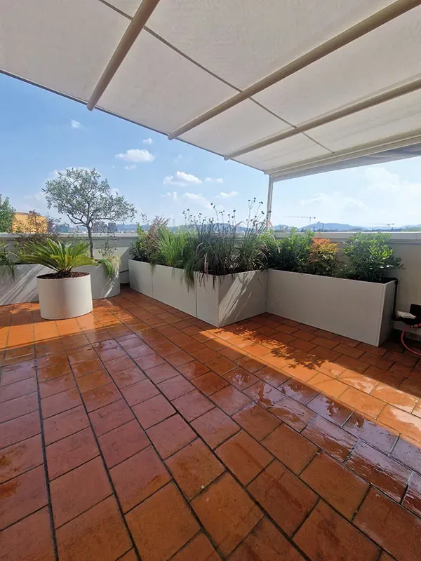 Terrazza con pavimento in cotto bagnato, tenda da sole e fioriere chiare di graminacee e fiori lungo il parapetto