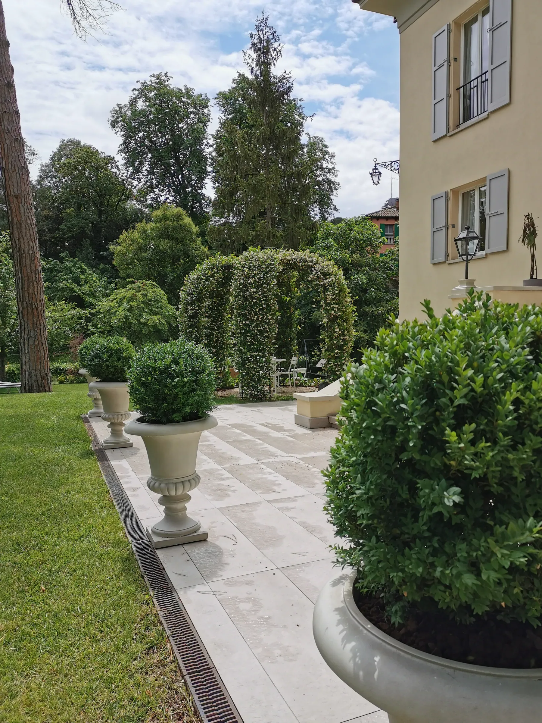 Viale in lastre di pietra chiara lungo il prato, con vasi di bosso a sfera e archi di rampicanti accanto a una villa